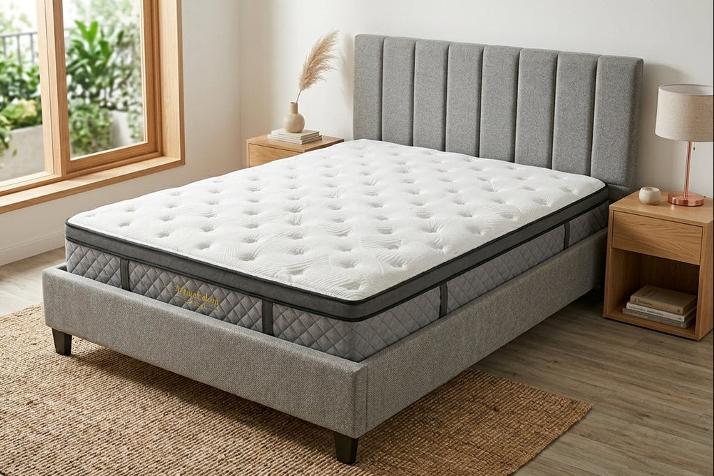 A white tufted mattress on a gray upholstered bed frame with a matching headboard, shown in a bright bedroom setting with wooden nightstands, a window, and a rug.