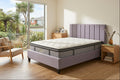 A bed with a tufted white mattress and a lavender upholstered frame and headboard, shown in a bright bedroom with wood floors, a chair, and a large window overlooking greenery.