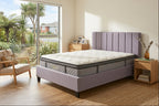 A bed with a tufted white mattress and a lavender upholstered frame and headboard, shown in a bright bedroom with wood floors, a chair, and a large window overlooking greenery.