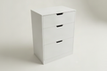 A three-drawer wooden nightstand or cabinet in a white finish, featuring recessed pull handles, photographed on a light neutral studio background.
