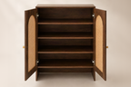 A dark brown wooden storage cabinet with two doors swung open, showing four interior shelves and woven cane panel inserts on the door faces, photographed against a warm beige studio background.