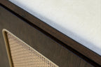 Close-up detail of a modern furniture piece featuring a dark wood cabinet base, a beige woven cane panel, and a light cream upholstered top surface.