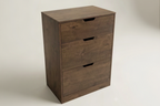 A three-drawer wooden nightstand or cabinet in a medium-to-dark brown finish, featuring recessed pull handles, photographed on a light neutral studio background.