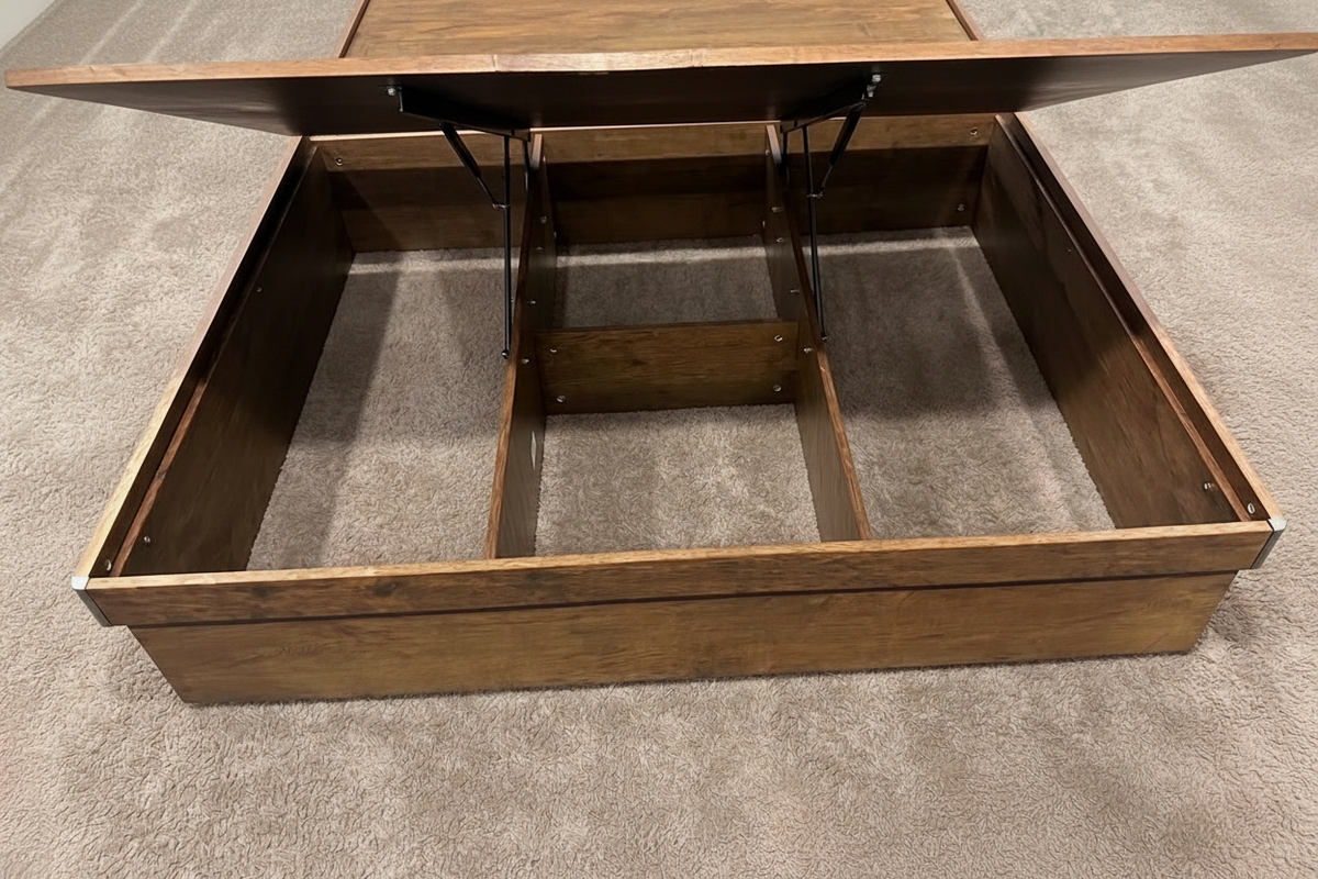 A dark brown wooden lift-top storage table shown open, revealing its raised top and internal compartment dividers, set on a gray carpeted floor.