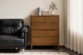 A wooden five-drawer chest of drawers shown in a styled interior scene next to a black leather sofa.