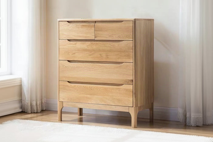 A light natural wood chest of drawers with five drawers and curved drawer pulls, shown in a minimalist room setting with curtains and a white wall.