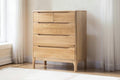 A light natural wood chest of drawers with five drawers and curved drawer pulls, shown in a minimalist room setting with curtains and a white wall.