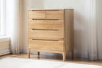 A light natural wood chest of drawers with five drawers and curved drawer pulls, shown in a minimalist room setting with curtains and a white wall.