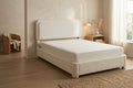 A modern upholstered bed frame in a warm cream bouclé fabric, shown in a styled bedroom setting with a matching mattress, a wooden nightstand, and a window with sheer curtains.