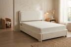 A modern upholstered bed frame in a warm cream bouclé fabric, shown in a styled bedroom setting with a matching mattress, a wooden nightstand, and a window with sheer curtains.