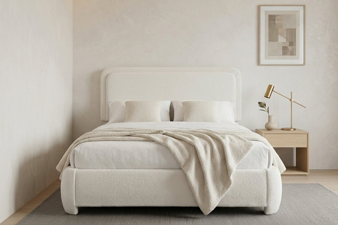 A modern upholstered bed in a minimalist bedroom setting, featuring a white fabric headboard and footboard, dressed in white bedding with a light beige throw blanket. A light wood nightstand with a gold desk lamp is positioned on the right against a textured beige wall with a framed abstract print.