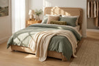 A styled bed in a bright bedroom featuring sage green bedding, a textured cream throw blanket, a beige upholstered headboard and footboard, and warm wood flooring.