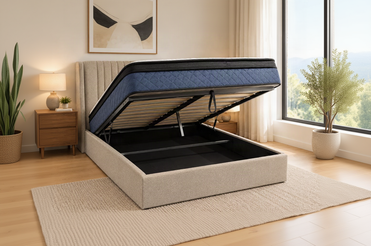 A beige upholstered lift-up storage bed with a raised mattress, shown in a bright bedroom setting with a window, bedside lamps, and a rug.