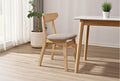 Wooden chair with gray cushion next to a wooden table in a room with a plant and curtains.