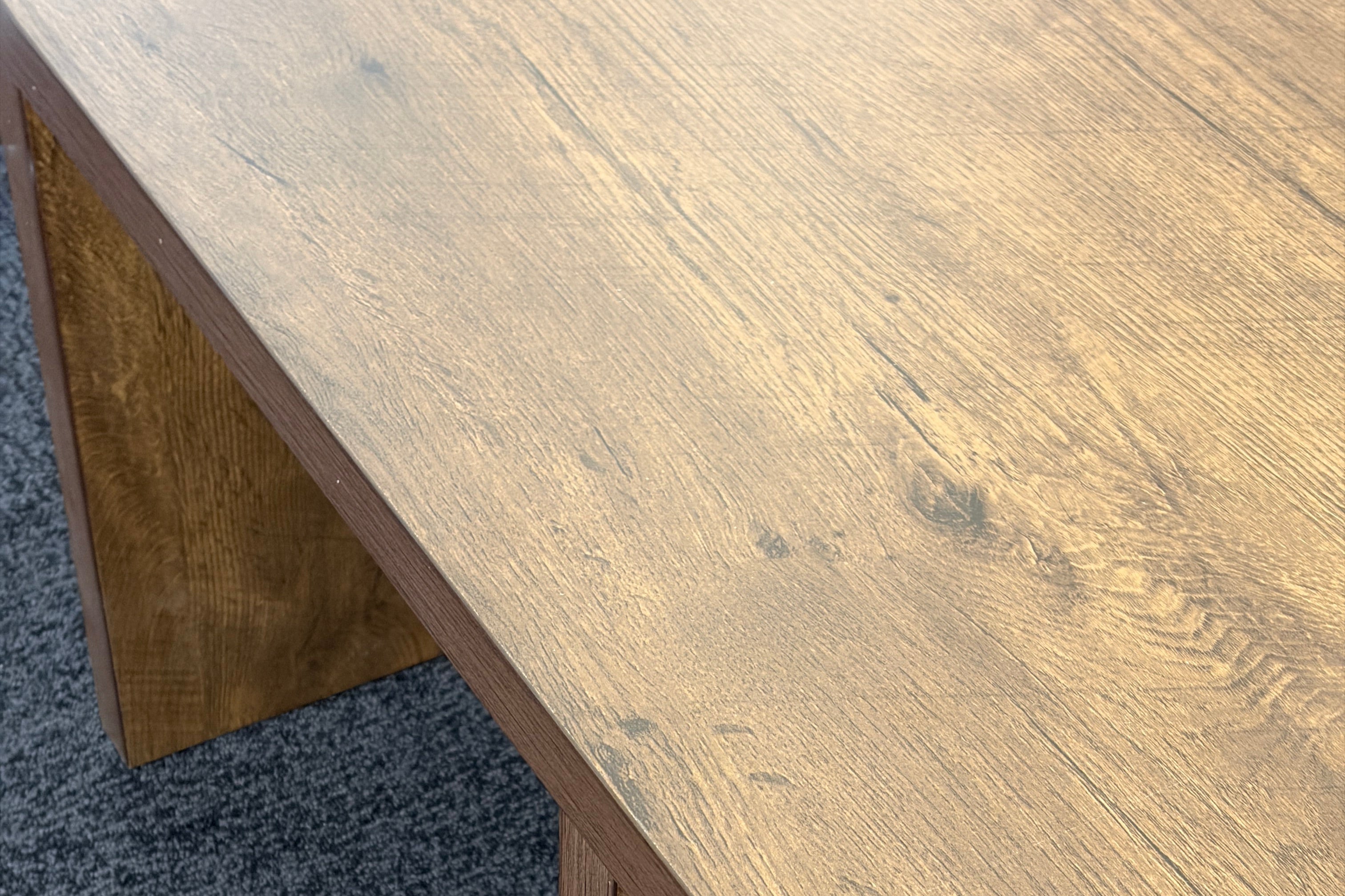 Close-up of a wooden table corner showing a medium brown wood grain tabletop and matching block-style legs on a dark gray carpet.