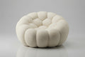 A round, low-profile pouf or accent chair upholstered in a textured cream fabric, featuring a segmented, flower-like petal design, set against a neutral gray background.