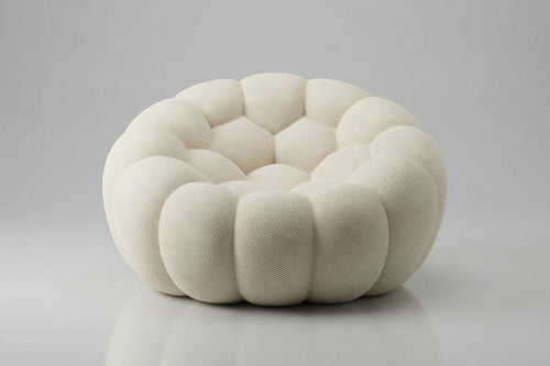 A round, low-profile pouf or accent chair upholstered in a textured cream fabric, featuring a segmented, flower-like petal design, set against a neutral gray background.